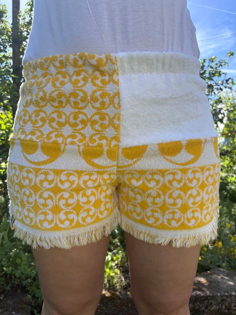 Vorderansicht der Frottee-Shorts Sole di Capri 73 im sonnengelben Retro-Design. Die Hose zeigt ein asymmetrisches Patchwork-Muster aus strahlend weißem Frottee und auffälligen, gelb-weißen Vintage-Ornamenten sowie verspielten Fransen am unteren Saum.