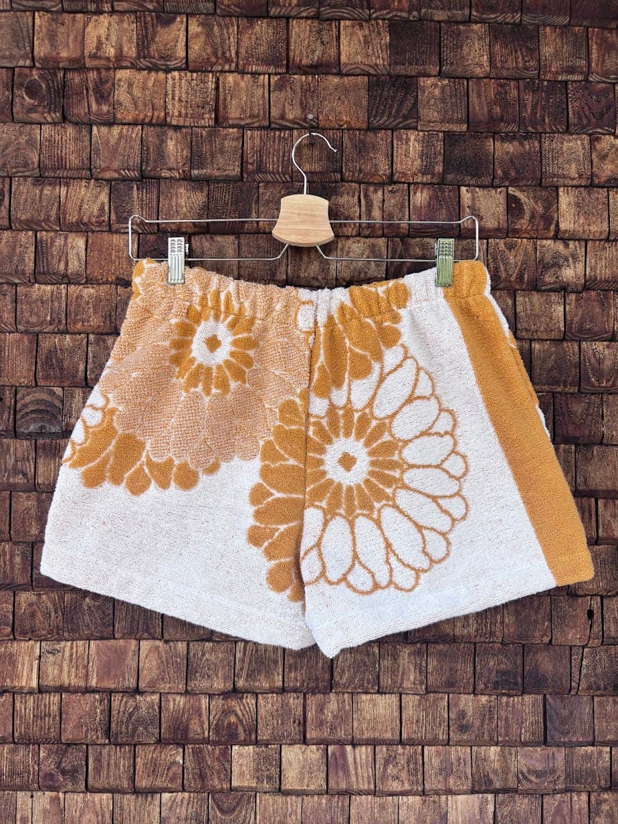 Rückansicht der Frottee-Shorts Sun Bloom 70 vor einer rustikalen Wandstruktur. Die Rückseite führt das weiße Frotteedesign mit den ockergelben 70s-Blütenmotiven fort und schließt auf der rechten Seite mit einem farblich abgesetzten, gelben Längsstreifen ab.