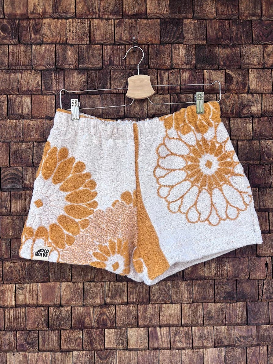 Vorderansicht der Upcycling-Frottee-Shorts Sun Bloom 70, hängend vor einer braunen Ziegelwand. Das asymmetrische Patchwork-Design zeigt große, florale Retro-Blütenmotive in einem warmen Ockergelb auf weißem Vintage-Frotteestoff mit dem eingenähten Old Wave Label am linken Saum.