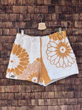 Vorderansicht der Upcycling-Frottee-Shorts Sun Bloom 70, hängend vor einer braunen Ziegelwand. Das asymmetrische Patchwork-Design zeigt große, florale Retro-Blütenmotive in einem warmen Ockergelb auf weißem Vintage-Frotteestoff mit dem eingenähten Old Wave Label am linken Saum.