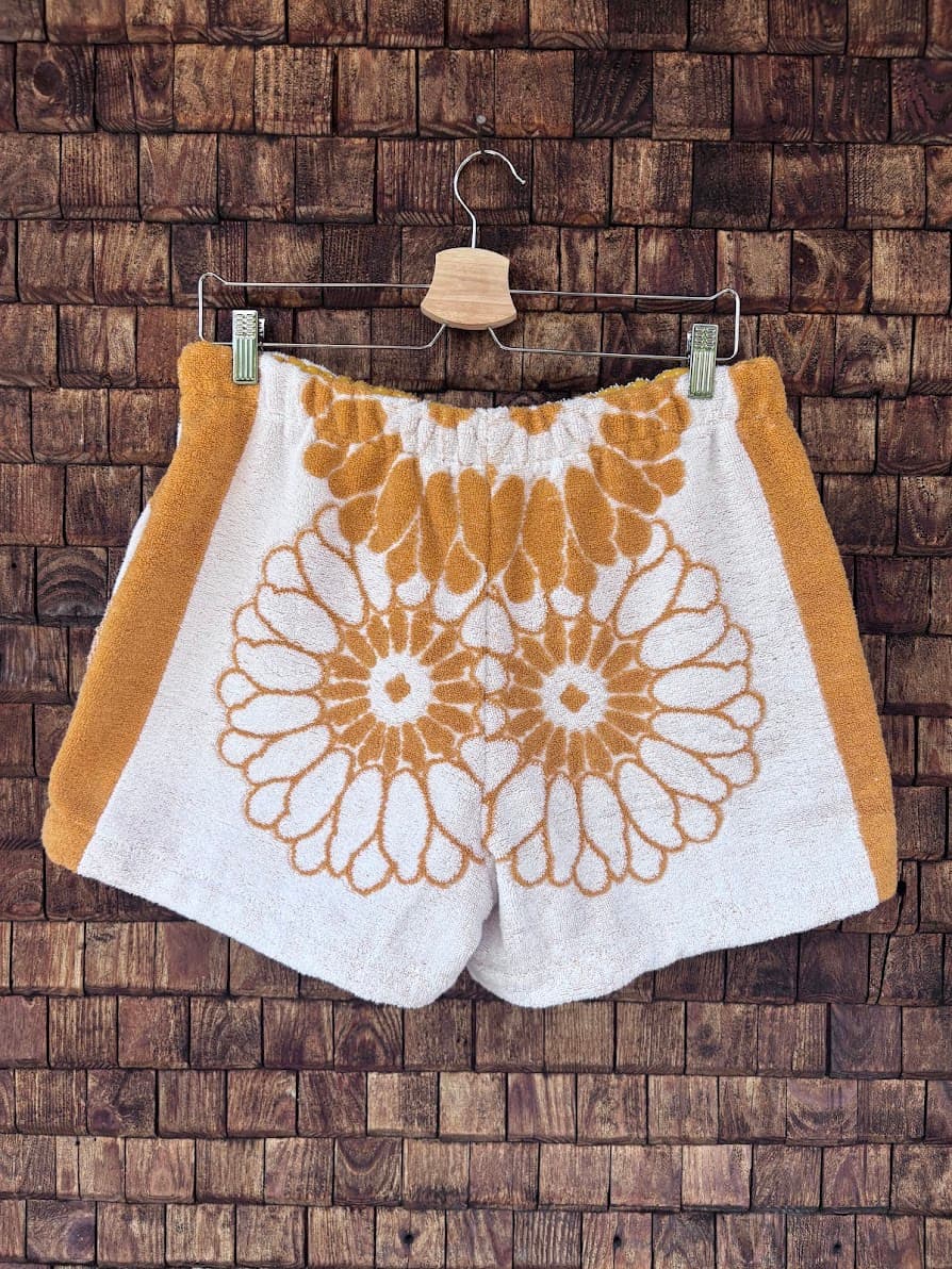 Rückansicht der Frottee-Shorts Sunfade Girasole 67 vor einem dunklen Holzhintergrund. Die Rückseite präsentiert sich mit einem markanten, spiegelbildlich angeordneten Sonnenblumen-Muster in Ockergelb auf weißem Grund sowie farblich abgesetzten, gelben Seitenstreifen.