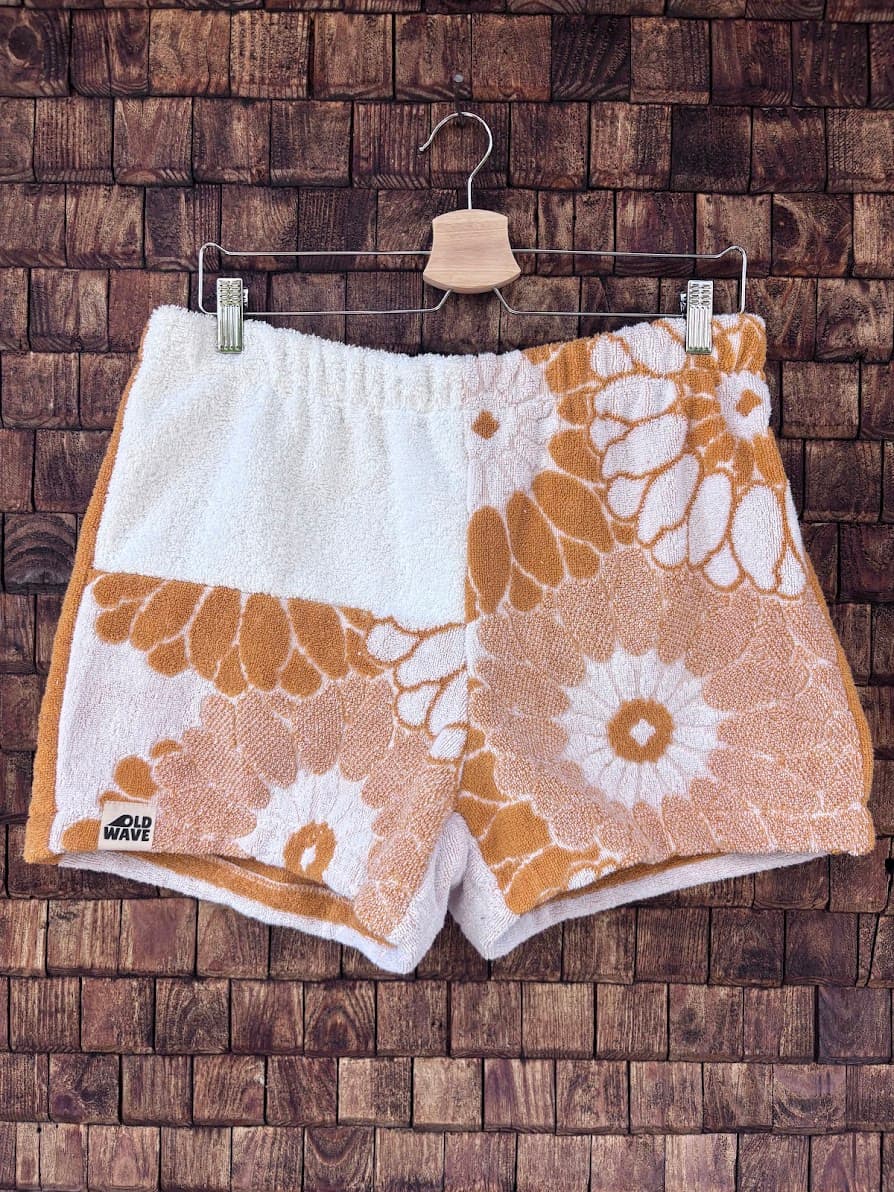 Vorderansicht der sommerlichen Upcycling-Frottee-Shorts Sunfade Girasole 67, hängend vor einer rustikalen Holzwand. Das asymmetrische Patchwork-Design besticht durch weiße Frotteestoffe, die mit großen, retro-inspirierten Sonnenblumen- und Blütenmotiven in einem warmen Ockergelb verziert sind, abgerundet durch das Old Wave Label am Saum.