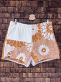 Vorderansicht der sommerlichen Upcycling-Frottee-Shorts Sunfade Girasole 67, hängend vor einer rustikalen Holzwand. Das asymmetrische Patchwork-Design besticht durch weiße Frotteestoffe, die mit großen, retro-inspirierten Sonnenblumen- und Blütenmotiven in einem warmen Ockergelb verziert sind, abgerundet durch das Old Wave Label am Saum.