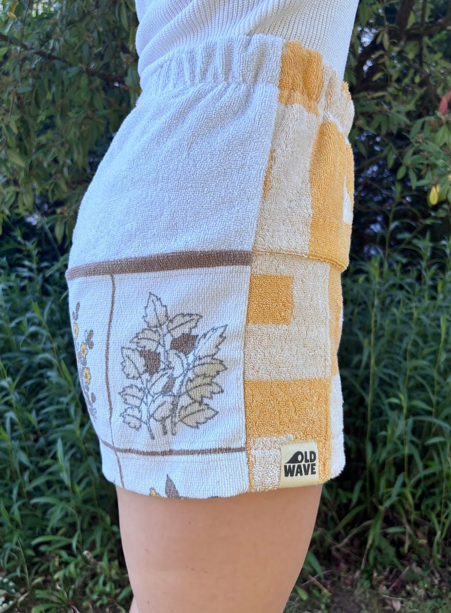 Seitliche Nahaufnahme der Shorts Sunfade Twist 71 am Modell. Der Fokus liegt auf der integrierten Hosentasche, dem weichen Vintage-Handtuchstoff und dem Übergang zum weiß-braunen Blumendruck.