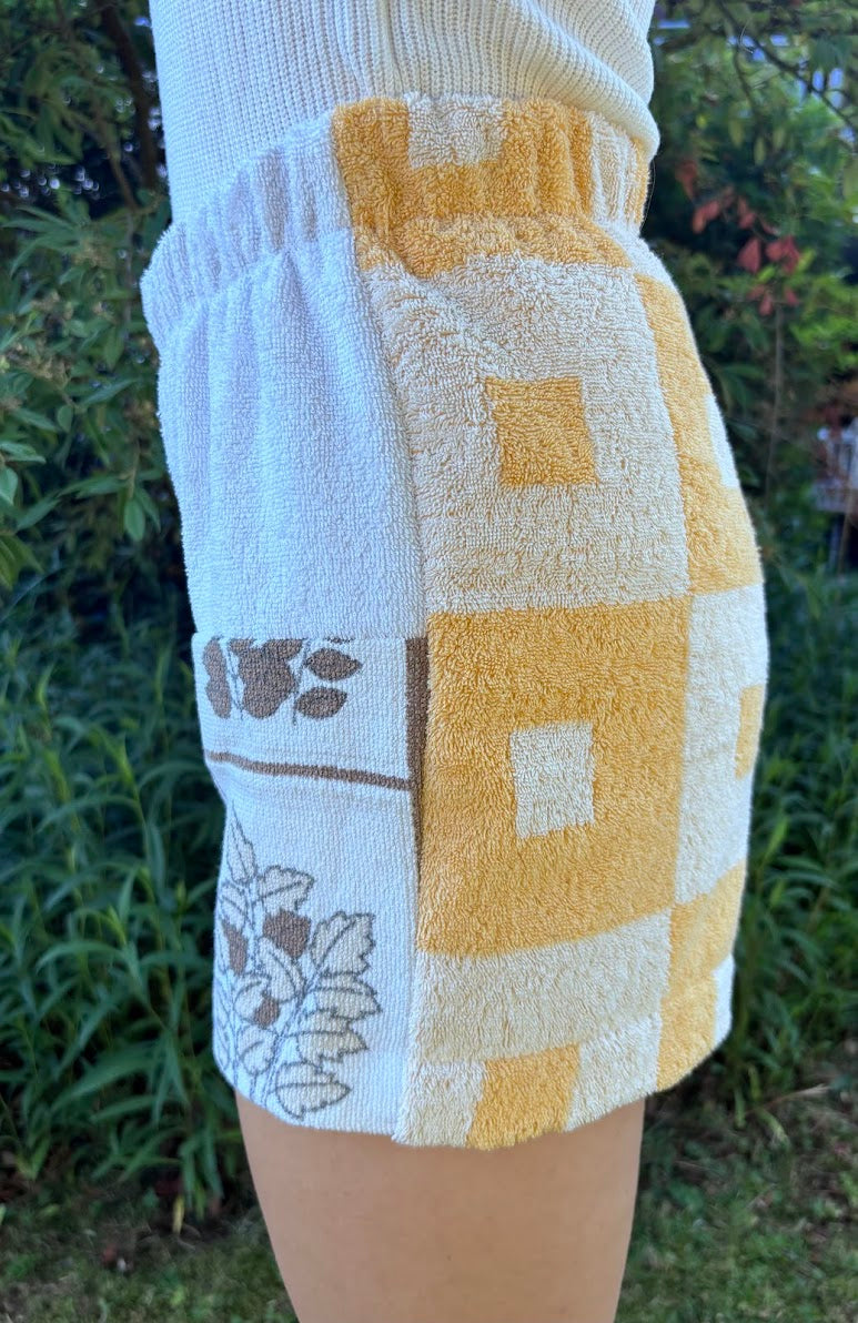 Seitliche Nahaufnahme der Shorts Sunfade Twist 71 am Modell. Der Fokus liegt auf der integrierten Hosentasche, dem weichen Vintage-Handtuchstoff und dem Übergang zum weiß-braunen Blumendruck.