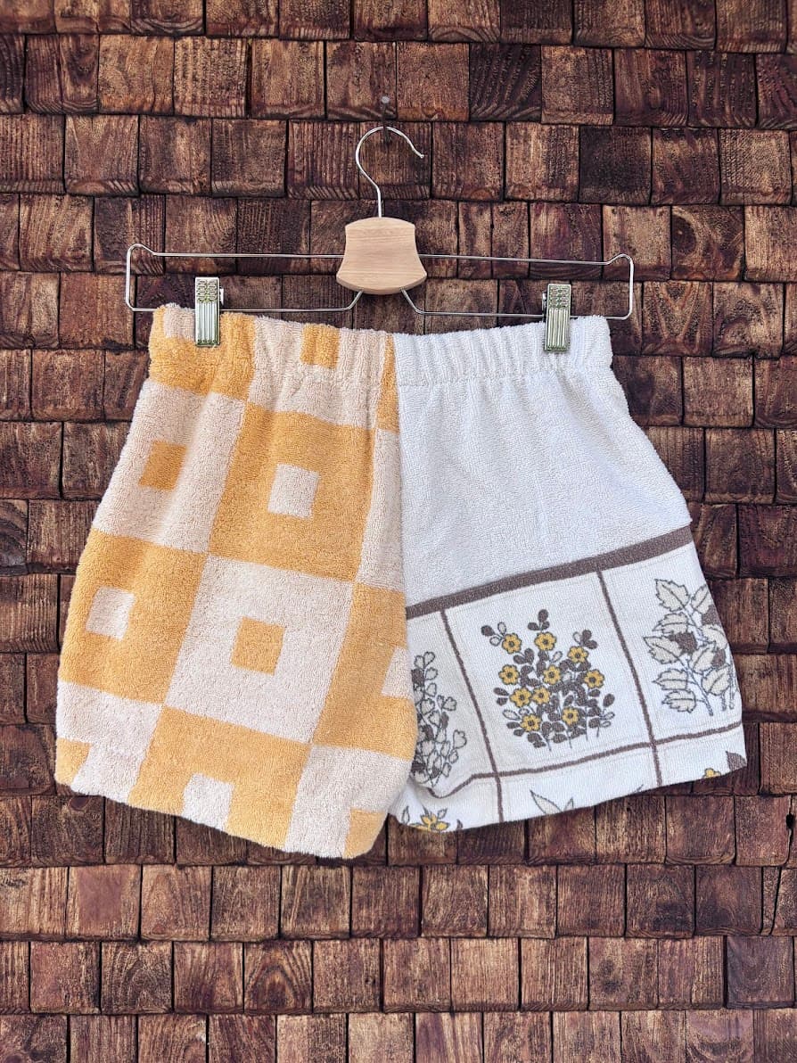 Rückansicht der Frottee-Shorts Sunfade Twist 71 vor einer dunklen Holzwand. Die Rückseite setzt das harmonische zweiteilige Design fort, bestehend aus der gelb-weiß karierten Stoffhälfte und der weißen Hälfte mit filigranen Retro-Blumenillustrationen.