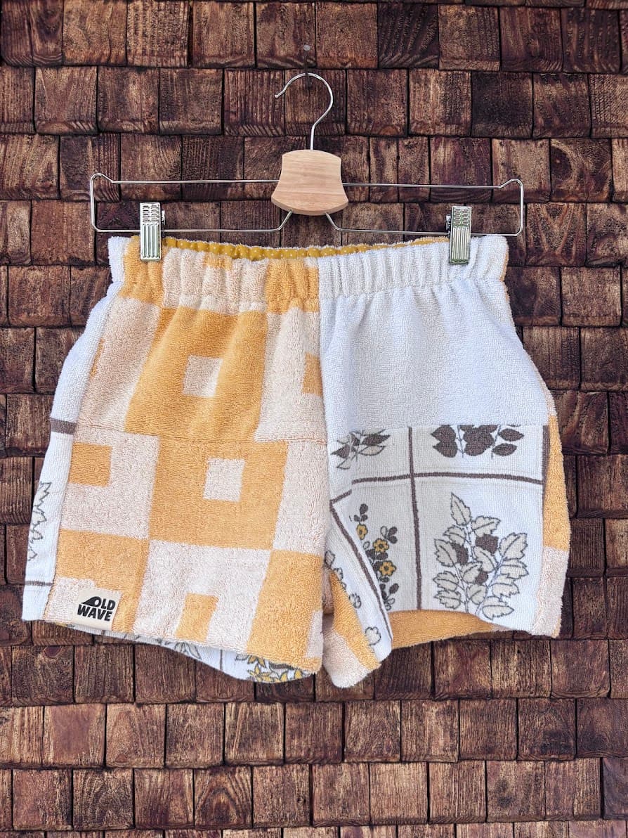 Vorderansicht der Upcycling-Frottee-Shorts Sunfade Twist 71, hängend vor einer rustikalen Holzwand. Das asymmetrische Patchwork-Design zeigt auf der linken Seite ein gelb-weißes Retro-Quadratmuster mit dem Old Wave Label am Saum und auf der rechten Seite weißen Vintage-Frotteestoff mit kunstvollen, braun skizzierten Blüten- und Kachelmotiven.