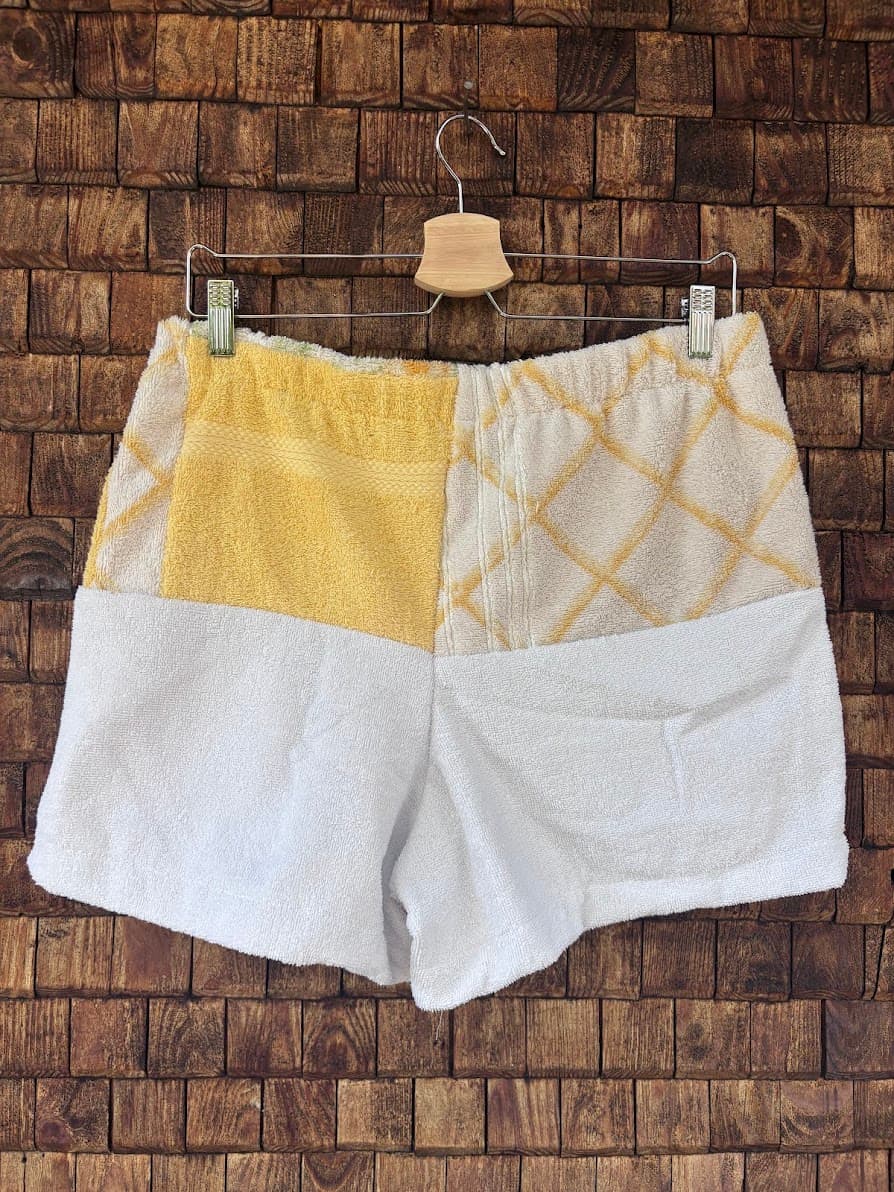 Rückansicht der Upcycling-Frottee-Shorts Terra Nocciola 66 vor einem dunklen Holzhintergrund. Die obere Hälfte der Shorts zeigt ein gelbes Retro-Muster sowie gelb-weiße Karostreifen, während die untere Hälfte komplett aus weichem, cleanem weißen Frotteestoff gefertigt ist.