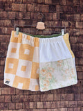 Vorderansicht der sommerlichen Upcycling-Frottee-Shorts Terra Nocciola 66, hängend vor einer rustikalen Holzwand. Das Patchwork-Design besticht auf der linken Seite durch ein gelb-weißes Retro-Quadratmuster mit dem Old Wave Label am Saum und auf der rechten Seite durch eine Kombination aus cleanem Weiß und einem zarten, floralen Vintagemuster.