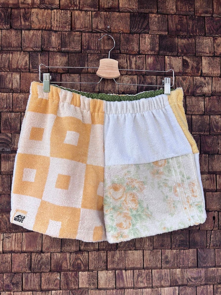 Vorderansicht der sommerlichen Upcycling-Frottee-Shorts Terra Nocciola 66, hängend vor einer rustikalen Holzwand. Das Patchwork-Design besticht auf der linken Seite durch ein gelb-weißes Retro-Quadratmuster mit dem Old Wave Label am Saum und auf der rechten Seite durch eine Kombination aus cleanem Weiß und einem zarten, floralen Vintagemuster.