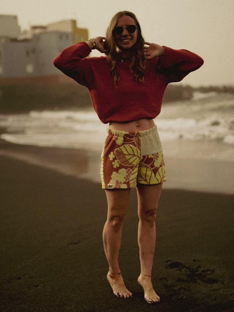 Ganzkörper-Lookbook-Aufnahme am dunklen Sandstrand bei bewölktem Himmel. Gezeigt wird das komplette Outfit aus den Terra Sunset 75 Frottee-Shorts, kombiniert mit einem langärmligen, weiten Crop-Strickpullover in Rostrot.