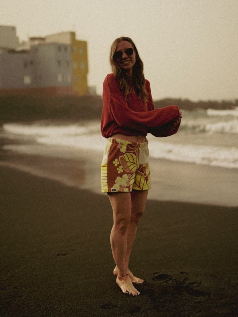 Seitliche Ansicht des Outfits am Strand in gedämpften, warmen Farbtönen. Das Model trägt die gemusterte Frottee-Hose und den weiten, herbstlich roten Pullover vor der Kulisse der Meereswellen.