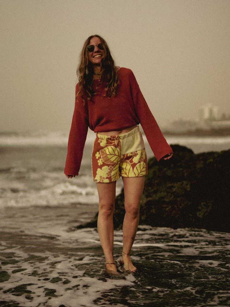 Stimmungsvolles Outdoor-Shooting im flachen, schäumenden Meerwasser. Frontale Ansicht des Outfits mit den Terra Sunset 75 Frottee-Shorts und dem rostfarbenen Pullover vor anrollenden Wellen.