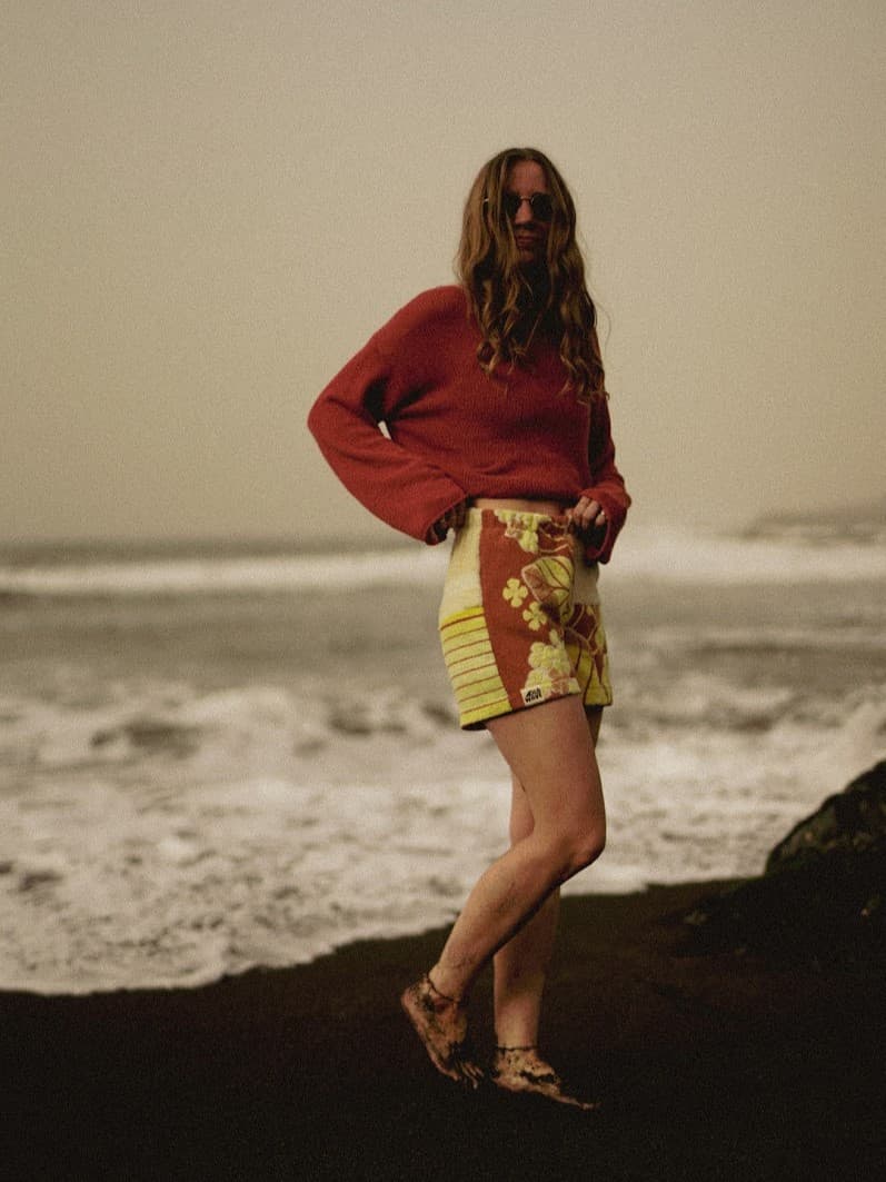 Profil-Aufnahme am dunklen Strand vor herannahender Gischt. Das herbstlich-warme Outfit aus Frottee-Shorts und weitem Crop-Pullover fügt sich harmonisch in die raue, atmosphärische Küstenlandschaft ein.