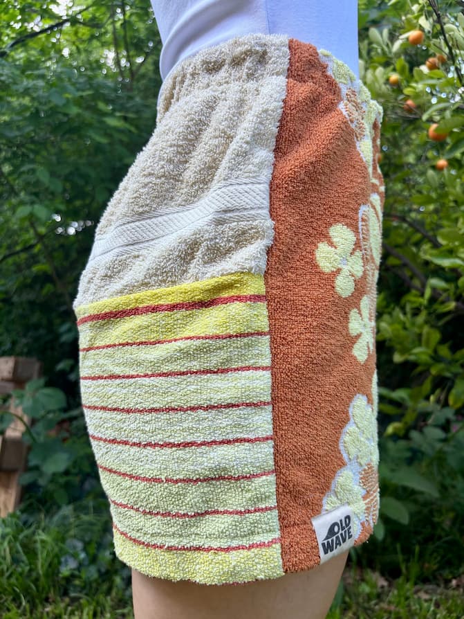 Getragene Seitenansicht von links. Zu sehen ist der Kontrast zwischen der terracottafarbenen Motivseite mit dem Old Wave Logo-Patch am Saum und der hinteren Beinhälfte, die aus hellgelbem Frottee mit dünnen, horizontalen Streifen in Terracotta besteht.