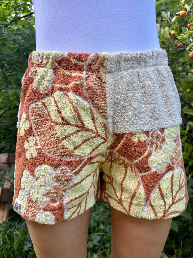 Getragene Vorderansicht der Terra Sunset 75 Shorts vor grünem Hintergrund. Die linke Seite zeigt ein großflächiges 70er-Jahre-Muster mit Blättern und Blüten in Terracotta und Gelb. Die rechte Seite hat im oberen Bereich ein sandbeiges Frottee-Inlay und darunter das florale Retromuster.