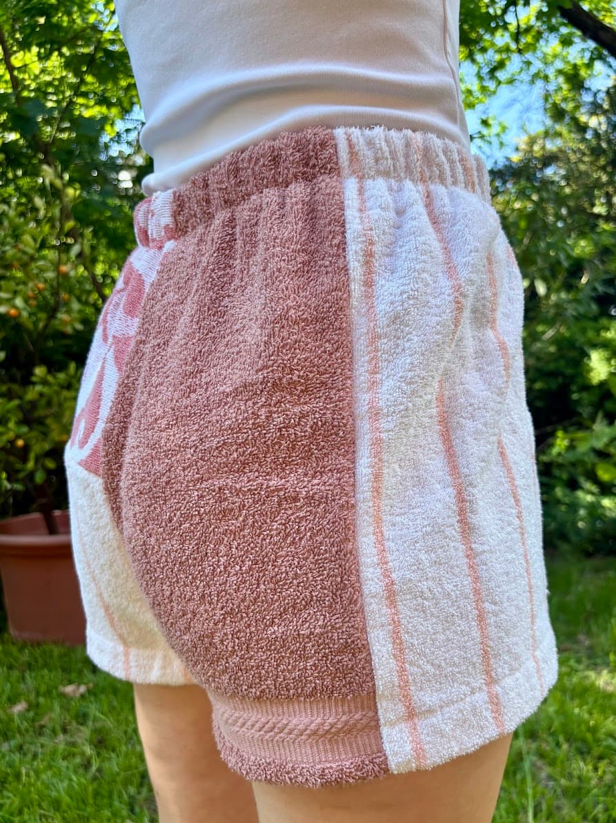 Getragene Rückansicht, schräg nach links gedreht. Zu sehen ist das linke Gesäß aus unifarbenem, siena-roséfarbenem Frottee, welches an der Außenseite in das kontrastierende weiße Frotteestück mit feinen Vintage-Streifen übergeht.