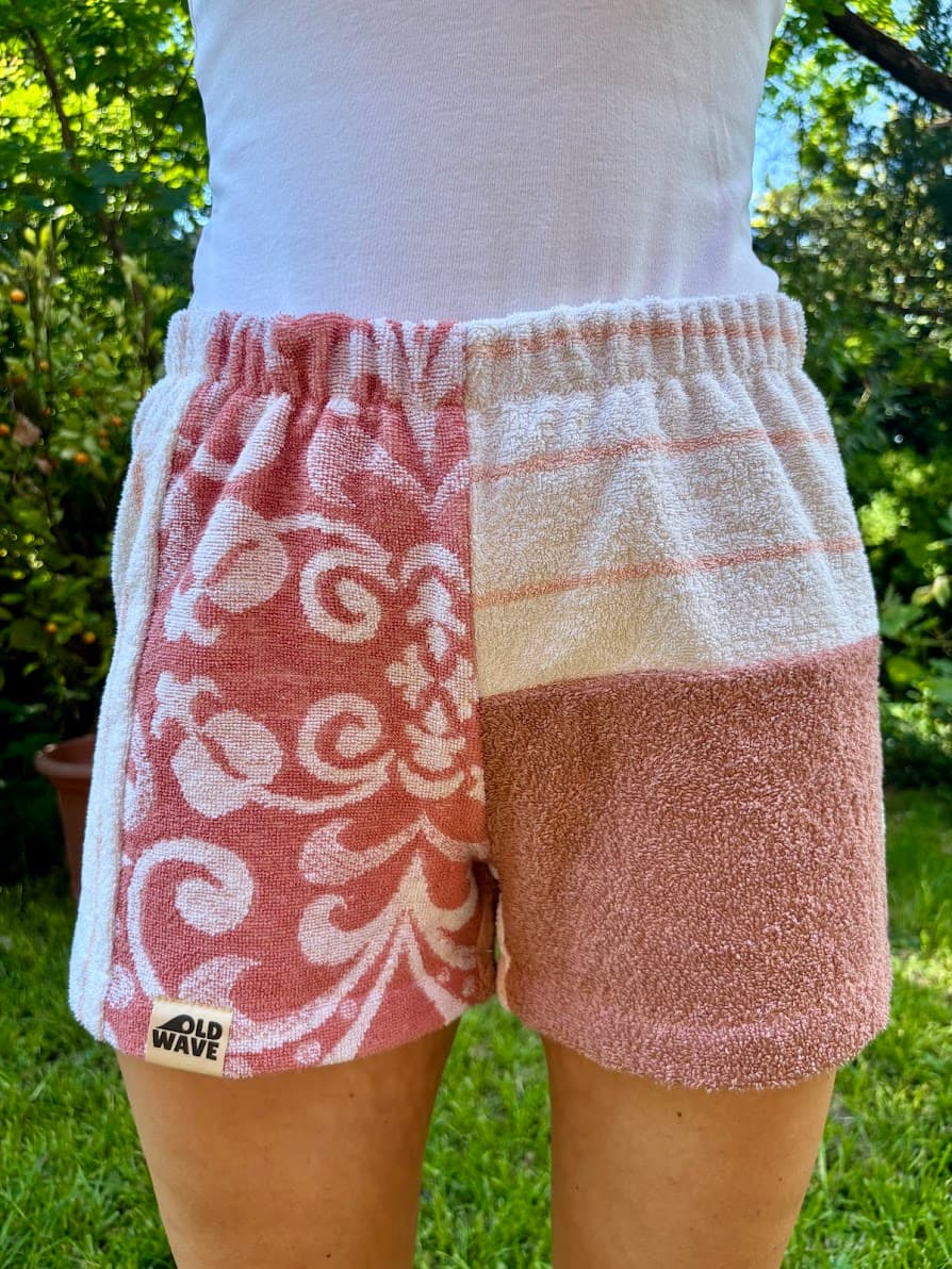 etragene Vorderansicht der Tramonto Siciliano 74 Shorts vor grünem Hintergrund. Das linke Bein zeigt barocke Ornamente in tiefem Rosé auf weißem Grund mit dem Logo-Patch am Saum. Das rechte Bein ist im oberen Teil weiß mit feinen horizontalen Linien in Siena-Orange, im unteren Teil unifarben siena-rosé.