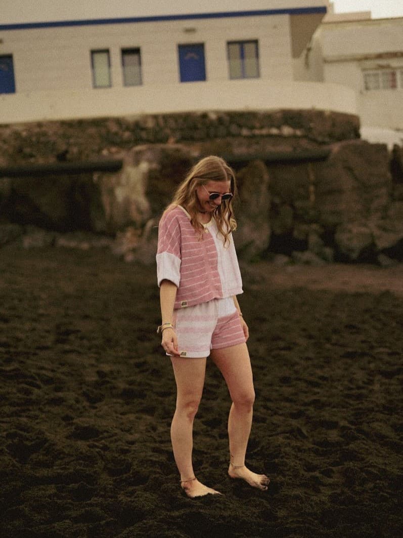 Outdoor-Lookbook-Foto auf dunklem Sandstrand. Gezeigt wird das lockere Towel Crop Shirt Blush Tide 77 aus weichem Vintage-Frottee im Half-and-Half-Design (eine Seite zartrosa gemustert, die andere weiß strukturiert), getragen als Matching-Set mit der passenden Frottee-Shorts.