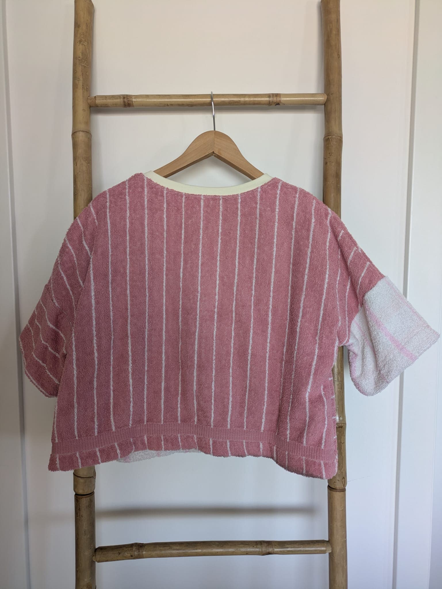 Rückansicht des Blush Tide 77 Towel Crop Shirts auf einem Kleiderbügel an einer Holzleiter. Die Rückseite spiegelt das Muster-Design wider: Der Hauptteil zeigt das weiße Vintage-Frottee mit den feinen rosa Längsstreifen, während der rechte Ärmelbereich kontrastierend aus dem roséfarbenen Ornament-Stoff angesetzt ist.