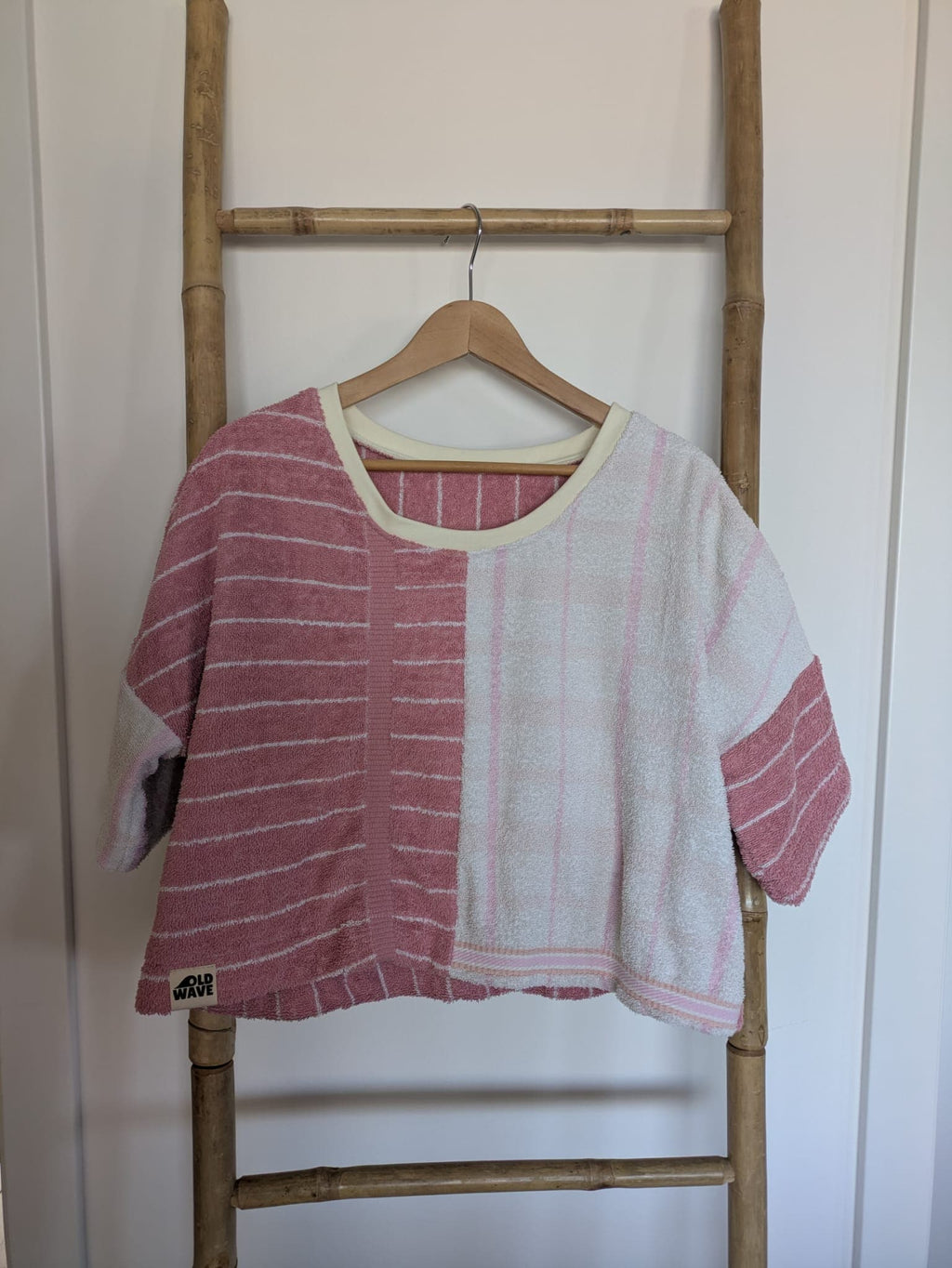 Vorderansicht des Blush Tide 77 Towel Crop Shirts, aufgehängt an einer rustikalen Holzleiter. Das lockere Oberteil besticht durch sein asymmetrisches Half-and-Half-Design aus Frottee: Die linke Seite zeigt florale Vintage-Ornamente in softem Rosé auf weißem Grund, während die rechte Seite aus weißem Frottee mit feinen, vertikalen rosa Streifen gefertigt ist.